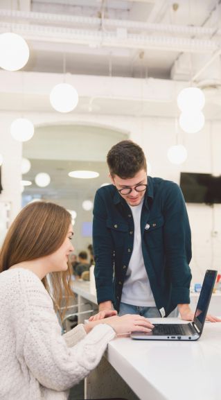 Two young adults collaborating over a laptop in a modern, bright office setting.
