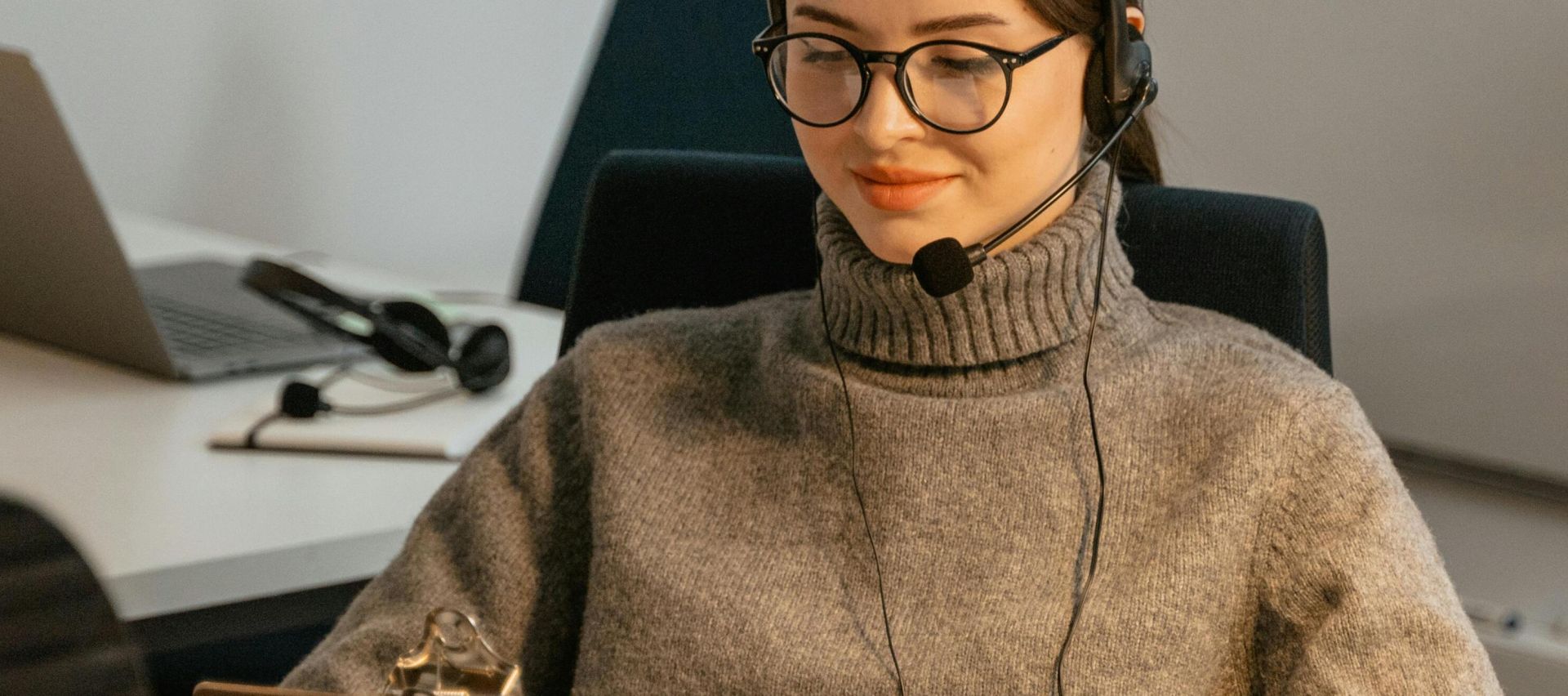 A smiling office worker with headphones, relaxed and reading a clipboard in a modern office.