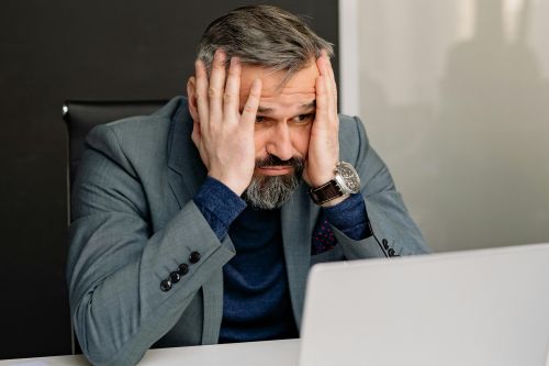 A stressed adult man in a suit at an office desk with hands on face and laptop.