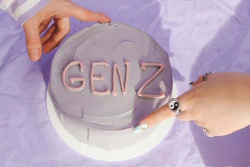 High angle view of a purple cake with 'Gen Z' icing touched by hands on a lavender surface.
