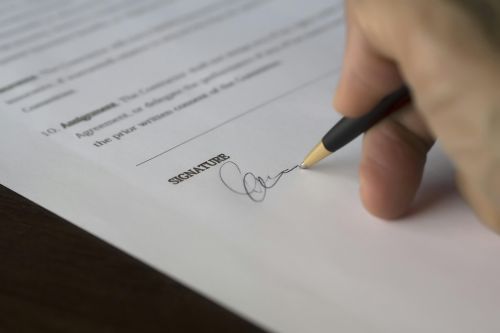 A hand signs a formal contract with a pen on a wooden desk.