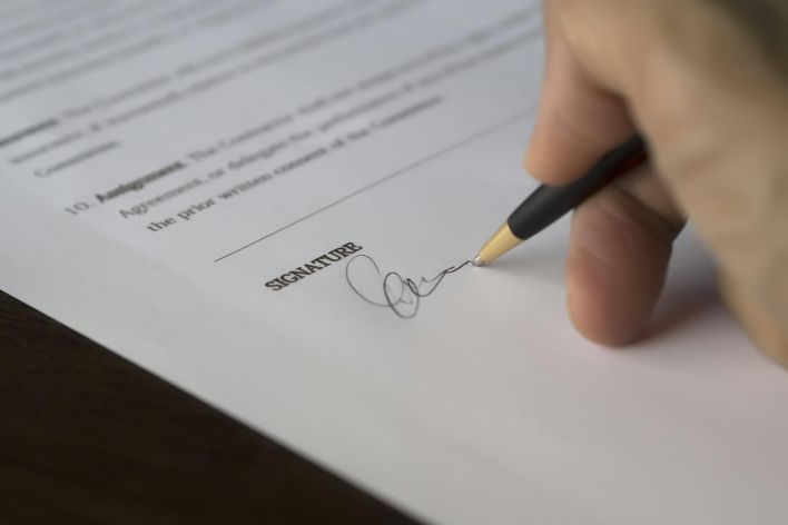 A hand signs a formal contract with a pen on a wooden desk.