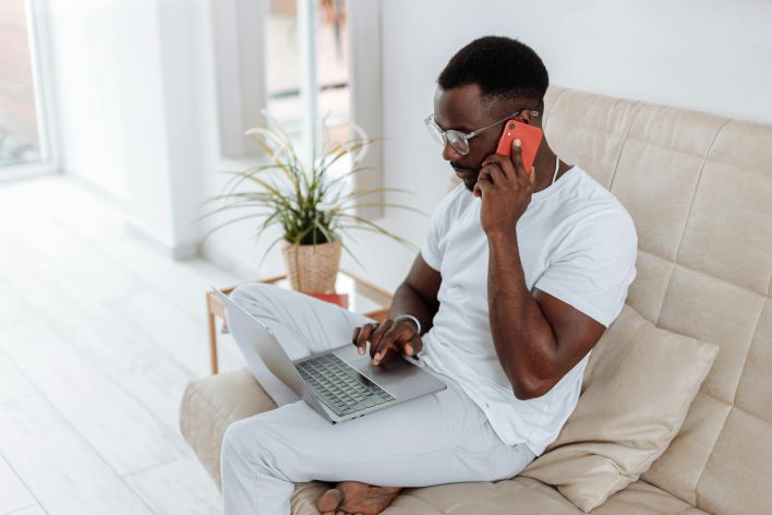 A man in a home office setting talking on the phone while working on a laptop.