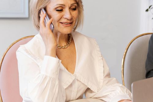 An elderly Caucasian woman multitasks on a laptop and smartphone in a modern office setting.